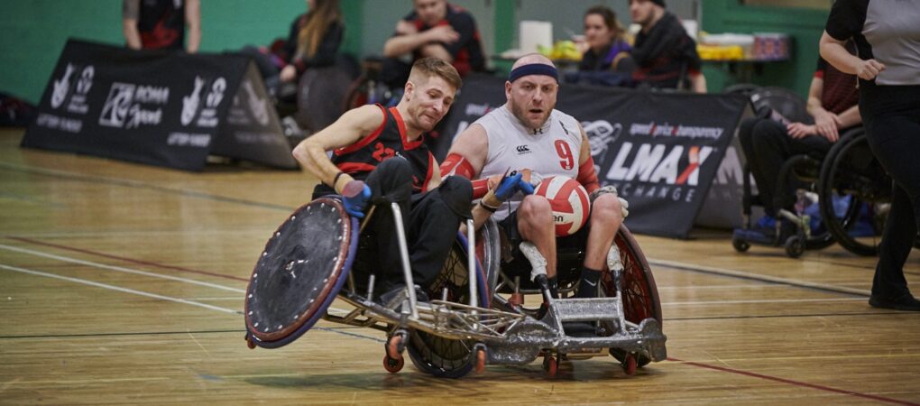 Wheelchair rugby player in black attempts to stop an opposing player who is wearing a black head band and white vest.