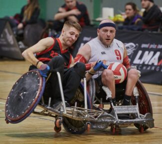 Wheelchair rugby player in black attempts to stop an opposing player who is wearing a black head band and white vest.
