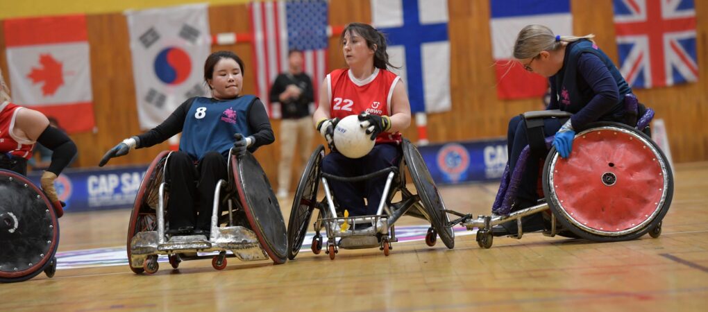 Female wheelchair rugby player in red vest advances up the court whilst being challenged by another female player.