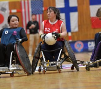 Female wheelchair rugby player in red vest advances up the court whilst being challenged by another female player.