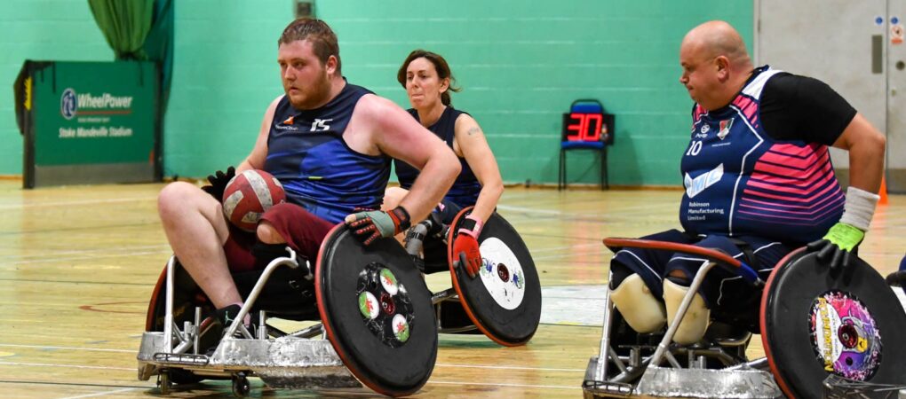 Berkshire Banshees player is pushing with the ball on his lap while being chased by a conventry stampede player.