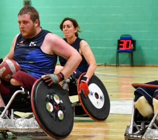 Berkshire Banshees player is pushing with the ball on his lap while being chased by a conventry stampede player.