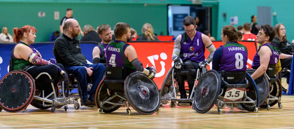 A team in purple vests called the 'Hawks' gather round to discuss team tactics during a game