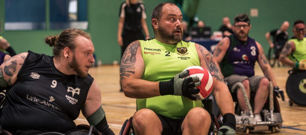 Northampton Saints player in a lime green vest is holding the red and white wheelchair rugby ball while under pressure from a defending player in a black vest,