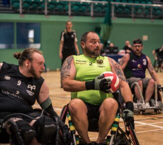 Northampton Saints player in a lime green vest is holding the red and white wheelchair rugby ball while under pressure from a defending player in a black vest,