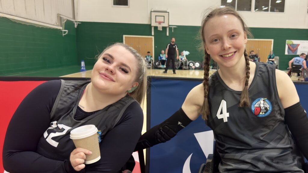 Two female wheelchair rugby players smiling to camera.