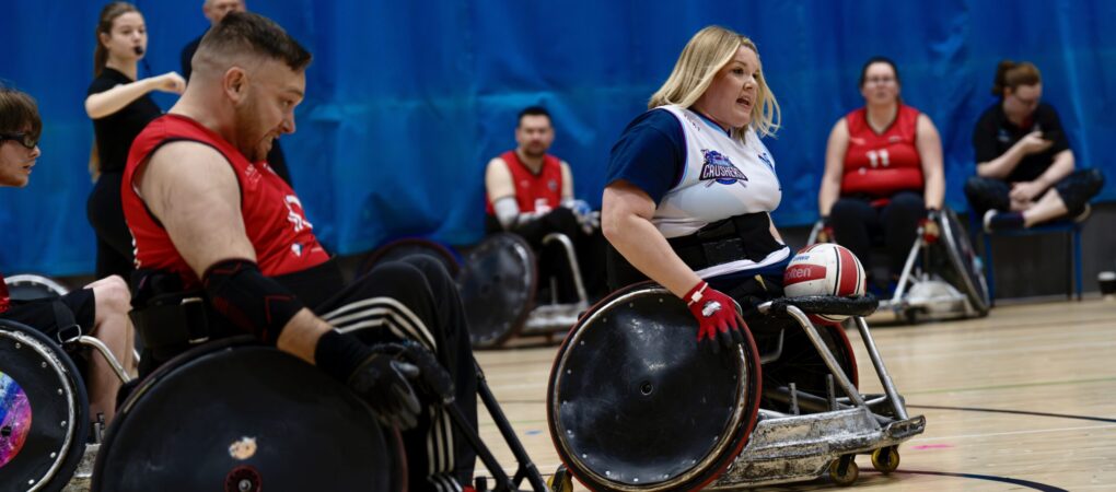 Female wheellchair rugby player carrying the ball. blonde hair, white and blue shirt, red and white ball.