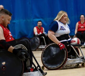 Female wheellchair rugby player carrying the ball. blonde hair, white and blue shirt, red and white ball.