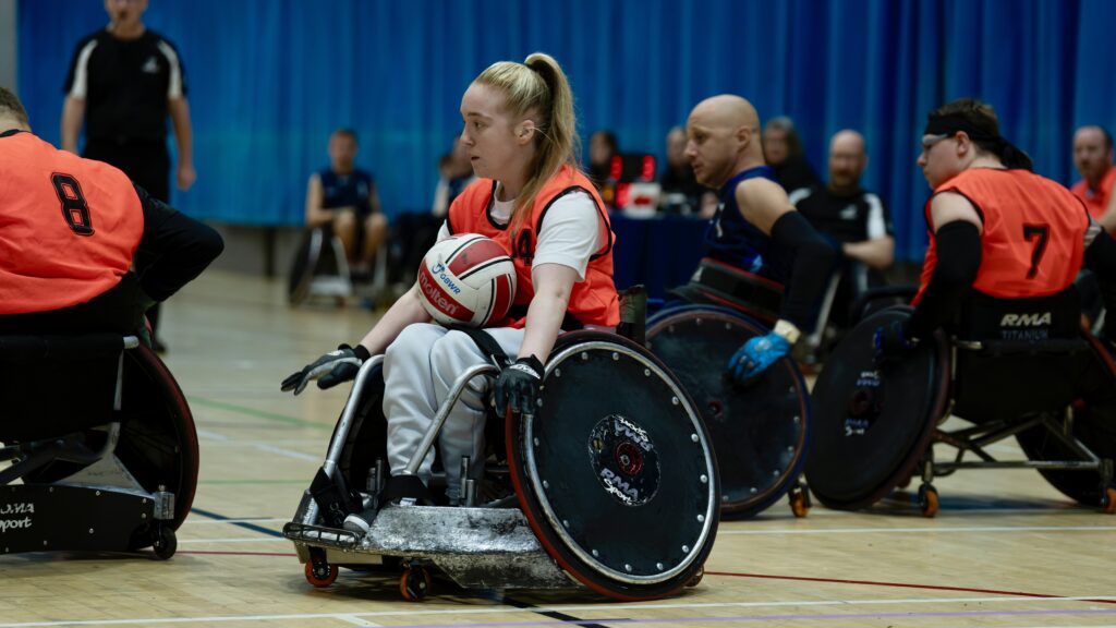 Female wheelchair rugby player wearing orange, pushes down court with a red and white ball.