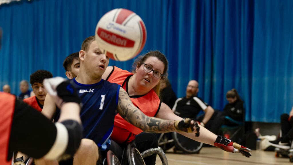 Two players trying to catch a rd and white ball playing wheelchair rugby