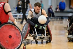 Male wheelchair rugby player leaning forward to ctch a white ball, wearing a white vest in a sports hall.