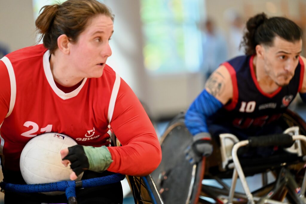 female wheelchair rugby player in a whiote vest protecting the ball from a male player wearing a blue vest.