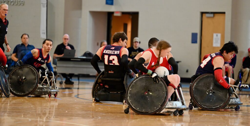 female wheelchair rugby player carrying the ball, wearing a red vest and blonde pig tails.