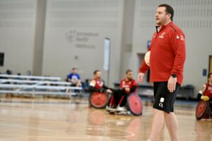 Lee Parry - Talent Head Coach carrying ball accross a wheelchair rugby court in america, wearing a red top and shorts.